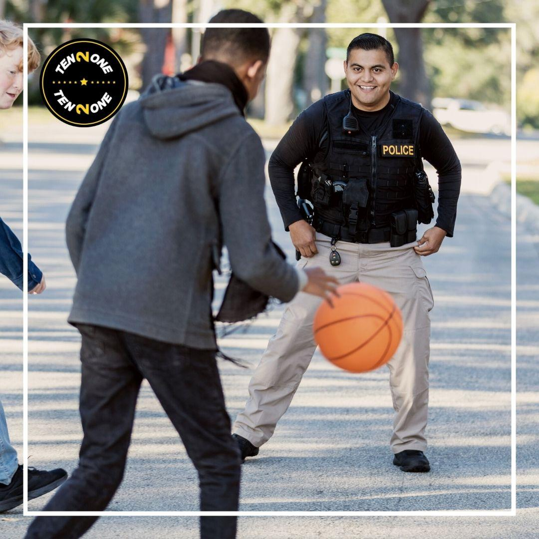 A person in a hoodie dribbles a basketball towards a police officer in tactical gear on a sunny street lined with trees