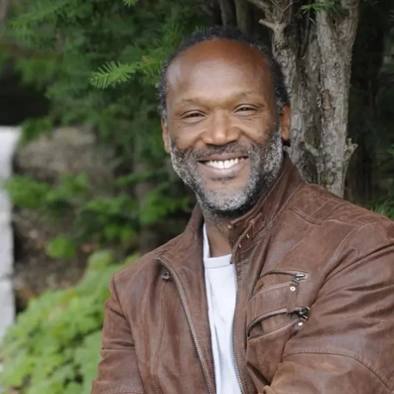 A person in a hoodie dribbles a basketball towards a police officer in tactical gear on a sunny street lined with trees
